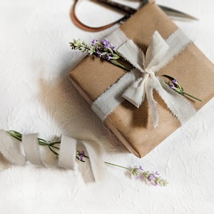 May include: A wrapped gift in brown paper, tied with a light gray ribbon and bow. Sprigs of lavender with purple flowers are tucked under the ribbon. Scissors are visible in the background, suggesting the gift is freshly wrapped.