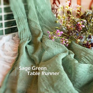 May include: A sage green table runner with a crinkled, textured fabric surface. The text "Sage Green Table Runner" is printed in white on the fabric. A small bouquet of dried wildflowers with pink, purple, and yellow petals rests on the runner.