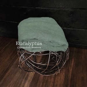 May include: A folded sage green cheesecloth runner with a loose, gauzy texture. The fabric is draped over a spherical, dark brown woven twig structure. The text "Eucalyptus Cheesecloth Runners" is printed in white on the fabric surface.