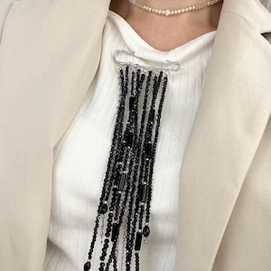 May include: A black beaded necklace with a large safety pin clasp. The necklace features strands of black beads and rectangular accents. A delicate pearl necklace is also visible. The person is wearing a white ribbed shirt and a beige blazer.