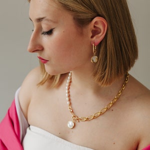 May include: A woman wearing a white strapless top and a pink jacket. She is wearing a gold chain necklace with a pearl pendant and matching gold earrings with a pearl pendant.