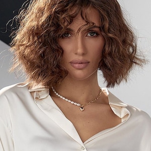 May include: A woman with brown curly hair wearing a cream-colored button-down shirt. A pearl necklace and a gold chain necklace with a heart pendant are around her neck. The background is a neutral color.
