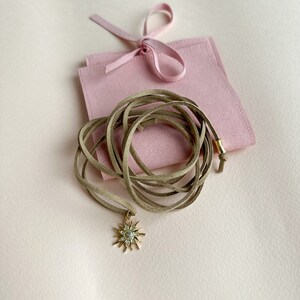 May include: A gold-colored sunburst pendant necklace with a tan suede cord. The necklace is displayed on a light pink surface next to a pink pouch tied with a ribbon. The pendant has a central stone surrounded by smaller stones.