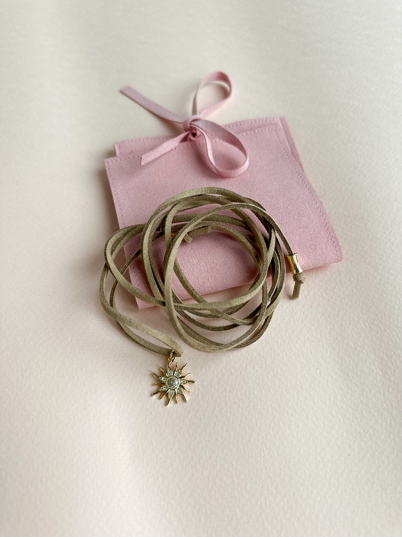 May include: A gold-colored sunburst pendant necklace with a tan suede cord. The necklace is displayed on a light pink surface next to a pink pouch tied with a ribbon. The pendant has a central stone surrounded by smaller stones.
