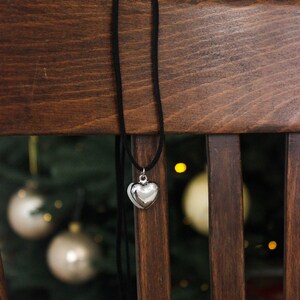 May include: A silver heart pendant necklace with a black cord hangs on a wooden chair. The heart-shaped pendant is shiny and reflective. The background features a Christmas tree with ornaments.