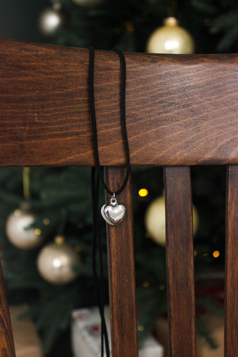 May include: A silver heart pendant necklace with a black cord hangs on a wooden chair. The heart-shaped pendant is shiny and reflective. The background features a Christmas tree with ornaments.