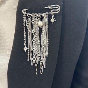 Könnte beinhalten: Eine silberne Ketten- und Stern-Anhänger-Brosche mit Perlenakzent. Die Brosche ist an einem schwarzen Blazer befestigt.