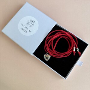 May include: A red necklace with a silver heart pendant is displayed inside a white box with a black interior. The box has the text "NATASSORY handmade jewelry designer" on a circular sticker. The necklace is made of a red cord.