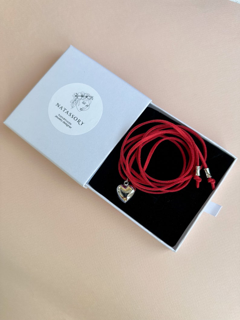 May include: A red necklace with a silver heart pendant is displayed inside a white box with a black interior. The box has the text "NATASSORY handmade jewelry designer" on a circular sticker. The necklace is made of a red cord.