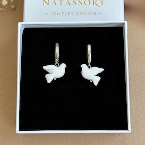 May include: A pair of white ceramic dove earrings with silver hoops. The earrings are in a black jewelry box with the text "NATASSORY - JEWELRY DESIGN -" on the lid.