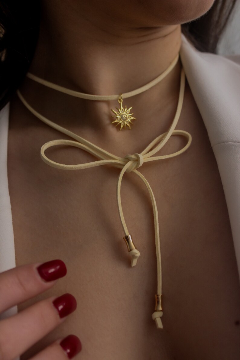 May include: A cream-colored choker necklace with a gold sun pendant. The necklace has a delicate, layered design with a bow detail. The sun pendant is detailed with a central stone and rays. The necklace is a stylish accessory.