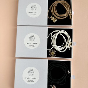 May include: Three white boxes with jewelry inside. Each box has a round sticker with the text "NATASSORY handmade jewelry designer". The jewelry includes a tan cord necklace with a gold charm, a white cord necklace with a gold sun charm, and a black cord necklace with a silver heart charm.