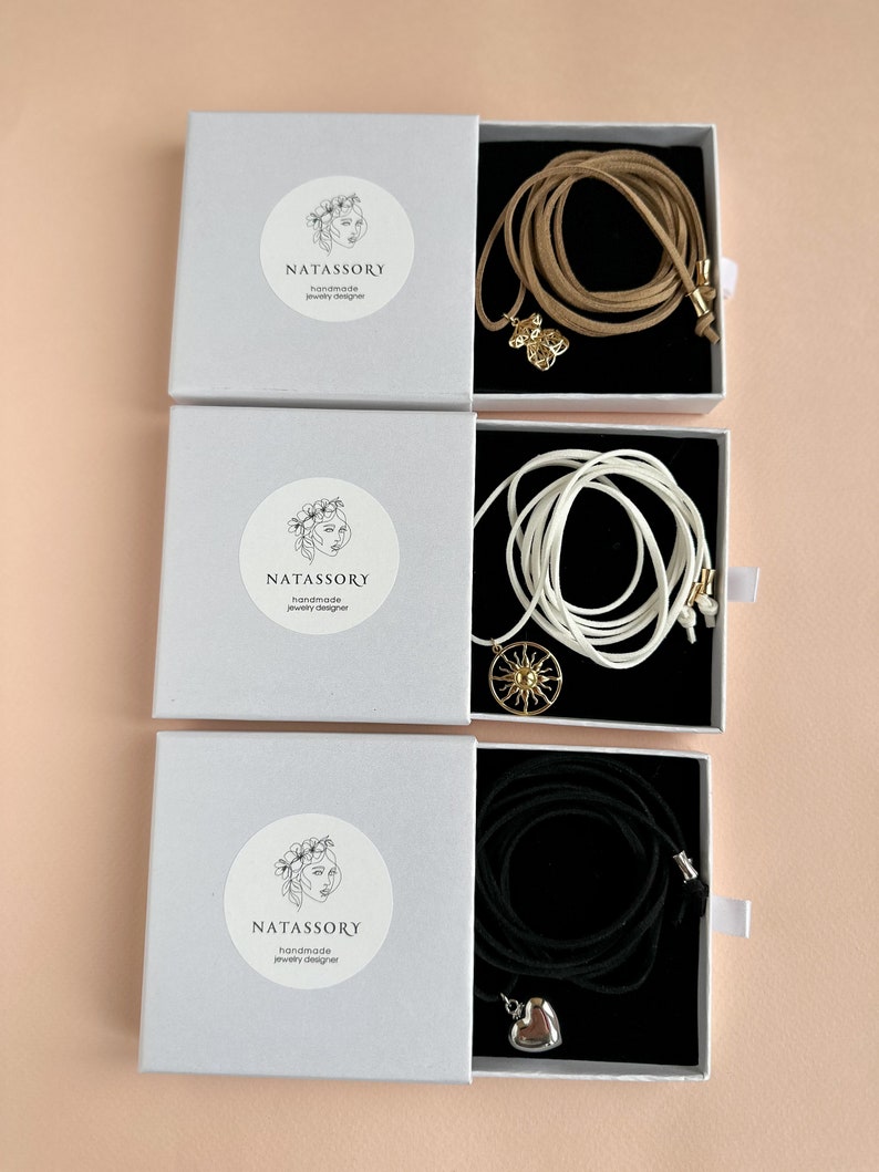 May include: Three white boxes with jewelry inside. Each box has a round sticker with the text "NATASSORY handmade jewelry designer". The jewelry includes a tan cord necklace with a gold charm, a white cord necklace with a gold sun charm, and a black cord necklace with a silver heart charm.