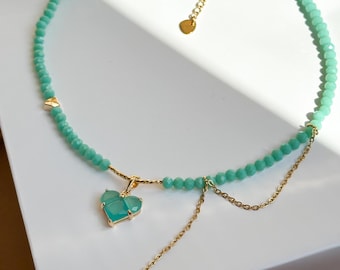 May include: A delicate necklace featuring a strand of small, light green beads. A gold-colored chain and a heart-shaped pendant with a light green stone add a touch of elegance. The necklace is displayed on a white surface.