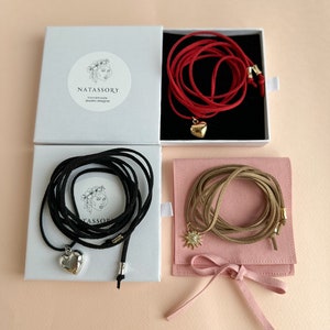 May include: Four necklaces in boxes and a pouch. One red necklace with a gold heart pendant, one black necklace with a silver heart pendant, and one tan necklace with a sunburst pendant. The boxes have the text "NATASSORY handmade jewelry designer".