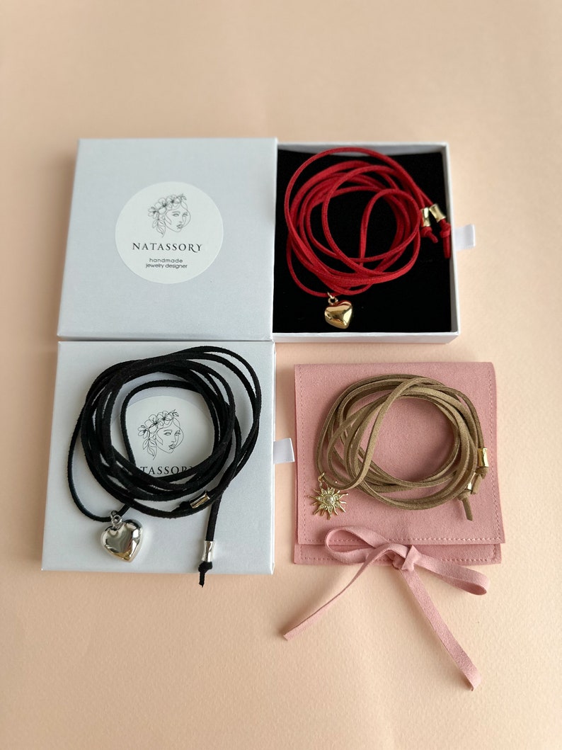 May include: Four necklaces in boxes and a pouch. One red necklace with a gold heart pendant, one black necklace with a silver heart pendant, and one tan necklace with a sunburst pendant. The boxes have the text "NATASSORY handmade jewelry designer".