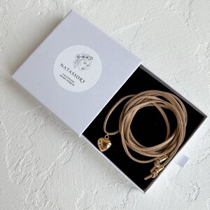 May include: A gold heart pendant necklace with a tan suede cord, presented in a white box with the brand name "NATASSORY" printed on it. The box is open, revealing the necklace against a black interior.