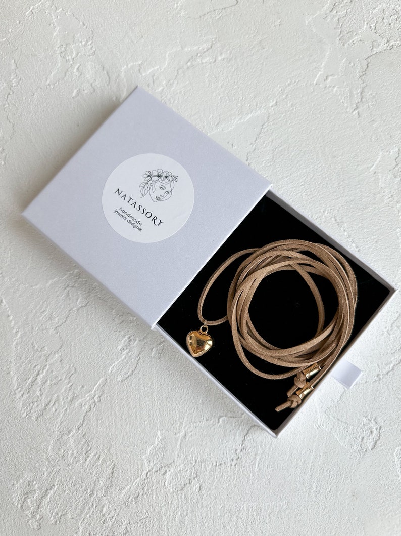 May include: A gold heart pendant necklace with a tan suede cord, presented in a white box with the brand name "NATASSORY" printed on it. The box is open, revealing the necklace against a black interior.