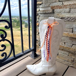 May include: A white western-style boot featuring a pointed toe, a low block heel, and decorative stitching on the shaft. A gold-toned ring is attached to the top pull tab, adorned with a hanging bow made of orange gingham and wavy purple ribbons.