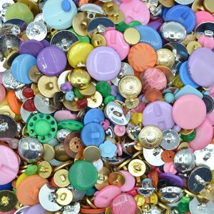 May include: A colourful collection of sewing buttons in diverse shapes and sizes. The assortment features round, flower, and heart-shaped buttons in pink, purple, blue, green, gold, and silver. A mix of plastic and metal buttons are visible.