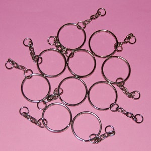 May include: A set of ten silver key rings with a chain attached to each ring. The key rings are arranged in a circle on a pink background.
