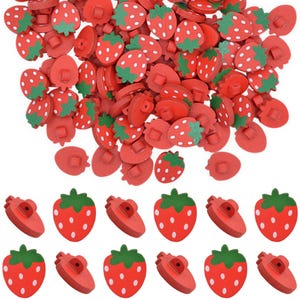 May include: A collection of small, strawberry-shaped buttons. Each button is painted red with white dots and features a green leaf top. The buttons are arranged in a loose pile at the top and in two organised rows at the bottom of the image.