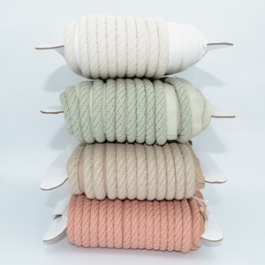 Pu&ograve; includere: Quattro bobine di corda intrecciata di diversi colori, impilate l'una sull'altra. Le bobine sono bianche, verde chiaro, beige e rosa.