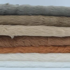 May include: A stack of six rectangular faux fur fabric swatches arranged in a vertical pile. The colors from top to bottom are dark gray, medium brown, light beige, medium tan, golden orange, and a base layer of white, all featuring a long pile.