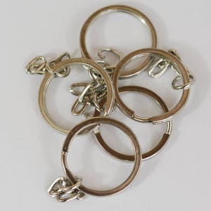 May include: A collection of silver key rings and chains, arranged in a slightly overlapping pattern against a white background. The key rings are circular, with a split ring design for attaching keys. The chains connect the rings.