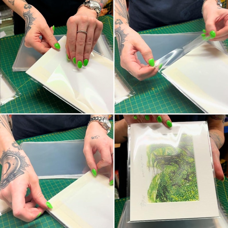 May include: A person is shown sealing a white matted print with a clear plastic sleeve. The print is of a green landscape with trees and a path. The person is using their fingers to smooth out the plastic sleeve.