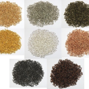 Op de afbeelding: Assortiment van kleine, metalen oogpins in verschillende kleuren, waaronder goud, zilver, brons, koper, zwart en antiek koper. De oogpins zijn gerangschikt in aparte stapels op een wit oppervlak.