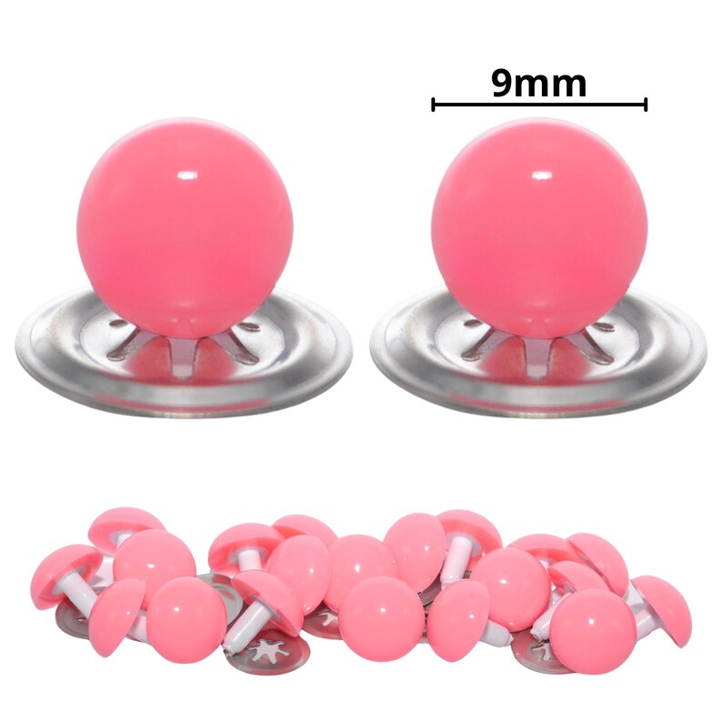Puede incluir: Un conjunto de ojos de seguridad de pl&aacute;stico rosa con acabado brillante, cada uno con una base de arandela de metal plateado. Dos de los ojos se muestran a mayor escala con una indicaci&oacute;n de medida de 9mm, mientras otros est&aacute;n dispersos.
