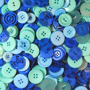 May include: A selection of blue and green buttons in various shades. The buttons have different sizes, shapes, and designs, including four-hole, two-hole, and patterned styles. The colour range spans from pale teal to dark navy.