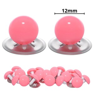 Puede incluir: Dos ojos de seguridad de pl&aacute;stico rosa grandes con arandelas de metal plateado sobre un mont&oacute;n de ojos de seguridad rosas m&aacute;s peque&ntilde;os a juego. El texto 12mm aparece sobre los ojos grandes, indicando un di&aacute;metro de 12mm para las piezas.