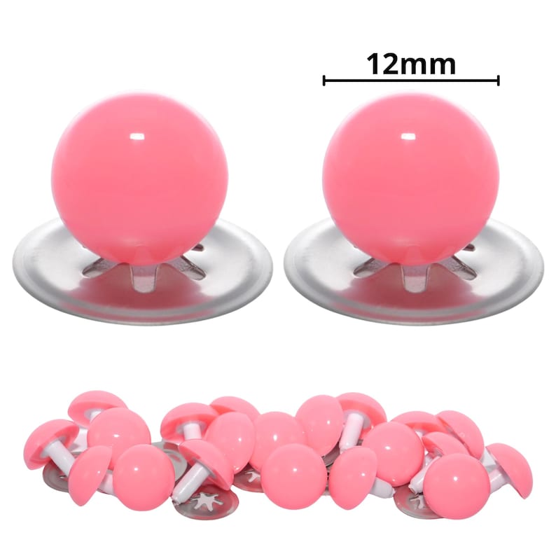 Puede incluir: Dos ojos de seguridad de pl&aacute;stico rosa grandes con arandelas de metal plateado sobre un mont&oacute;n de ojos de seguridad rosas m&aacute;s peque&ntilde;os a juego. El texto 12mm aparece sobre los ojos grandes, indicando un di&aacute;metro de 12mm para las piezas.