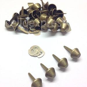 Könnte beinhalten: Eine Sammlung antiker, messingfarbener Polsternägel mit konischem Kopf und langem, schlankem Schaft. Mehrere Metallplatten sind ebenfalls sichtbar. Die Nägel sind auf einer weißen Oberfläche verstreut.