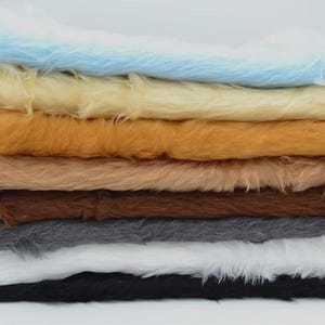 May include: A stack of rectangular faux fur fabric swatches in various colors including light blue, cream, golden yellow, tan, dark brown, gray, white, and black. Each piece features a long, soft, synthetic pile texture layered horizontally.