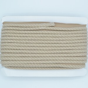 Pu&ograve; includere: Un rotolo di cordoncino di cotone beige con un'etichetta bianca. Il cordoncino &egrave; avvolto stretto e ha una texture sottile.