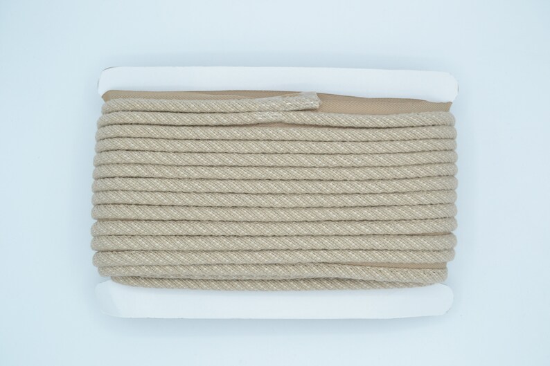 Pu&ograve; includere: Un rotolo di cordoncino di cotone beige con un'etichetta bianca. Il cordoncino &egrave; avvolto stretto e ha una texture sottile.