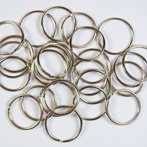 Könnte beinhalten: Eine Ansammlung von silberfarbenen Metall-Schlüsselringen, die auf einer weißen Oberfläche verstreut sind. Die Ringe sind rund und haben eine kleine Öffnung zum Befestigen von Schlüsseln oder anderen Gegenständen. Das Bild ist eine Nahaufnahme, die die Details der Ringe zeigt.