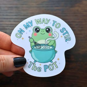 May include: A whimsical sticker featuring a green frog stirring a blue cauldron with a whisk. The text "ON MY WAY TO STIR THE POT" is arched around the frog. The frog has rosy cheeks and large eyes. The sticker is held by a hand.