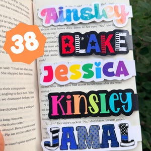 May include: Five rectangular bookmarks featuring the names Ainsley, Blake, Jessica, Kinsley, and Jamal in various fonts and colors. Ainsley has a watercolor effect, Blake features stars and stripes, Jessica is multicolored, Kinsley is neon, and Jamal has a blue line pattern.