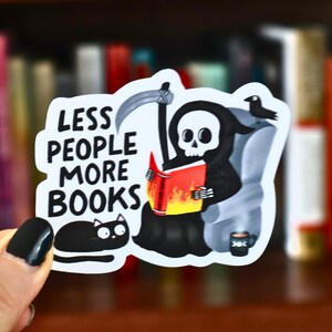 May include: A white sticker with the text "LESS PEOPLE MORE BOOKS" features a cartoon Grim Reaper reading a book with flames. A black cat and a coffee cup are also depicted. The sticker is held in front of a bookshelf.