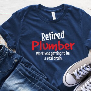 Peut inclure: Un t-shirt bleu marine avec le texte "Retired Plumber" en lettres blanches et rouges. Le texte "Work was getting to be a real drain." est imprimé en dessous en blanc.