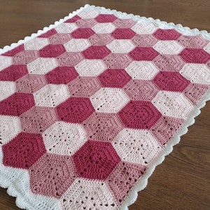May include: A crocheted baby blanket with a hexagonal pattern in shades of pink and white. The blanket has a white crocheted border.