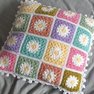 May include: A crocheted cushion with a daisy pattern in shades of pink, green, yellow, and purple. The cushion has a white crocheted border with pom-poms.