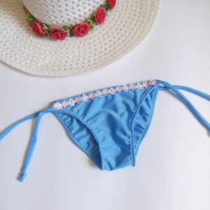Op de afbeelding: Een lichtblauwe bikinibroek met een roze en witte bloemenrand. De bikinibroek heeft aan elke kant verstelbare bandjes.