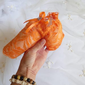 Op de afbeelding: Een feloranje kanten kousenband met een delicaat bloemenpatroon en een geschulpte rand. Het accessoire is versierd met een kleine, gladde oranje satijnen strik aan de bovenkant, waaraan een minuscuul zilverkleurig metalen bedeltje is bevestigd.