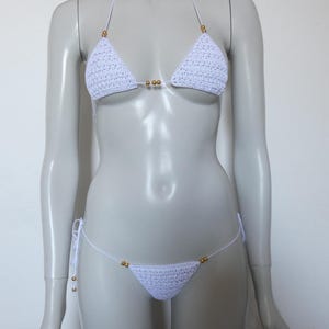 May include: A white crochet bikini set featuring triangular cups and a matching bottom. The design includes thin white string ties and small, round, metallic gold-colored beads positioned at the center of the top and along the sides of the bottom.
