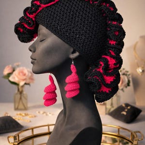 May include: A black crochet hat with a red trim and matching pink earrings are displayed on a mannequin. The hat has a ruffled edge, and the earrings are a stacked spiral design. The background includes a gold-rimmed tray and jewelry.
